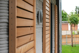Die Fassade aus dem 3D-Betondrucker wird durch Fensterläden und Giebel aus Holz ergänzt. Gauben und Biberschwanz-Dachziegel vervollständigen die klassische Anmutung des modernen Mehrfamilienhauses, das im KfW-55-Standard errichtet wurde.